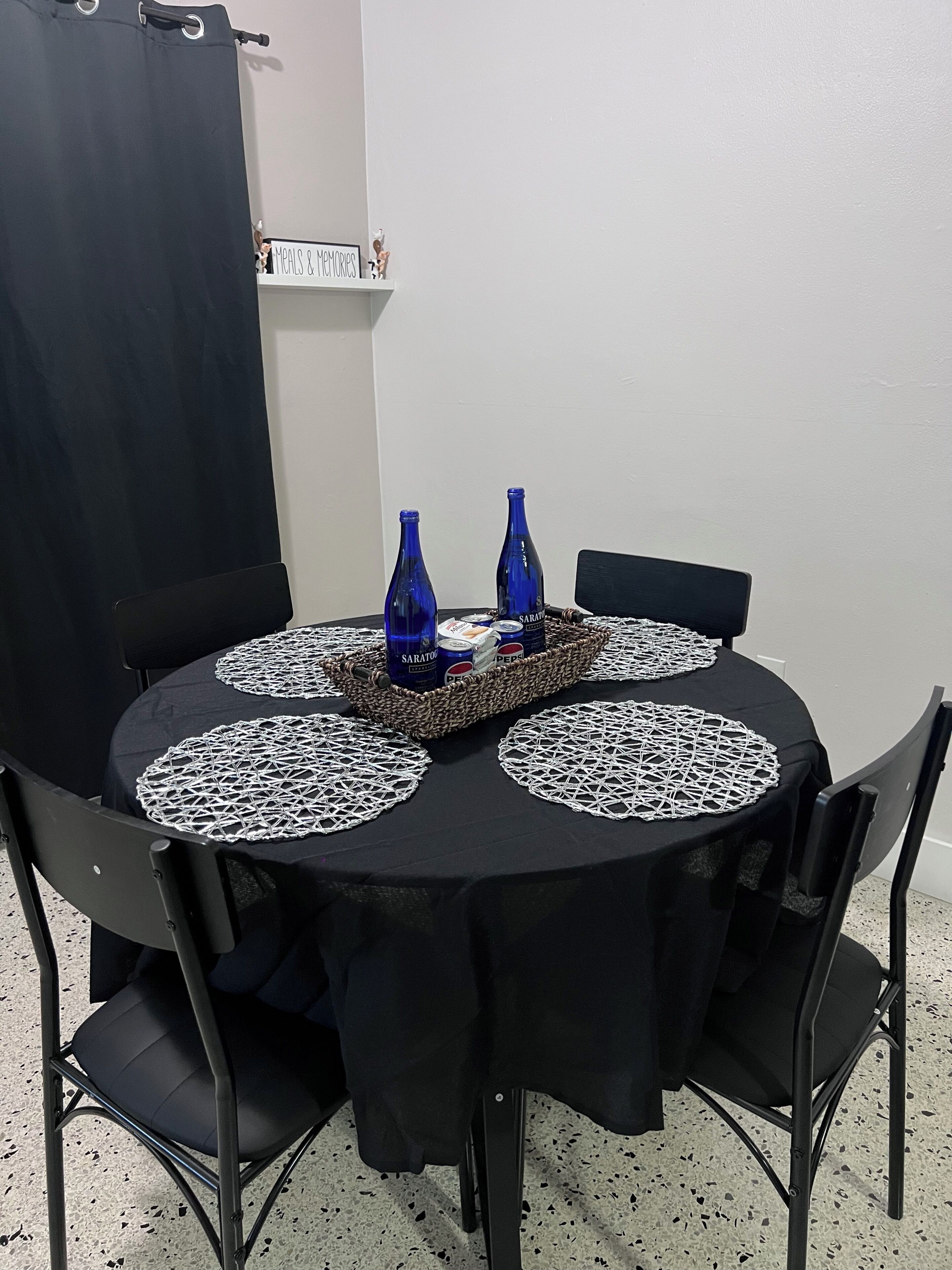 Une table ronde recouverte d’une nappe noire, entourée de quatre chaises, et deux bouteilles bleues posées sur la table.