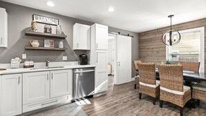 A modern kitchen with white cabinetry, a built-in dishwasher, and a dining area with wicker chairs. - 1 Almost Heaven (Panama City Beach)