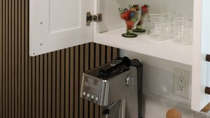 Et køkken med kaffemaskine, et skab med vinglas og en skål med frugt.