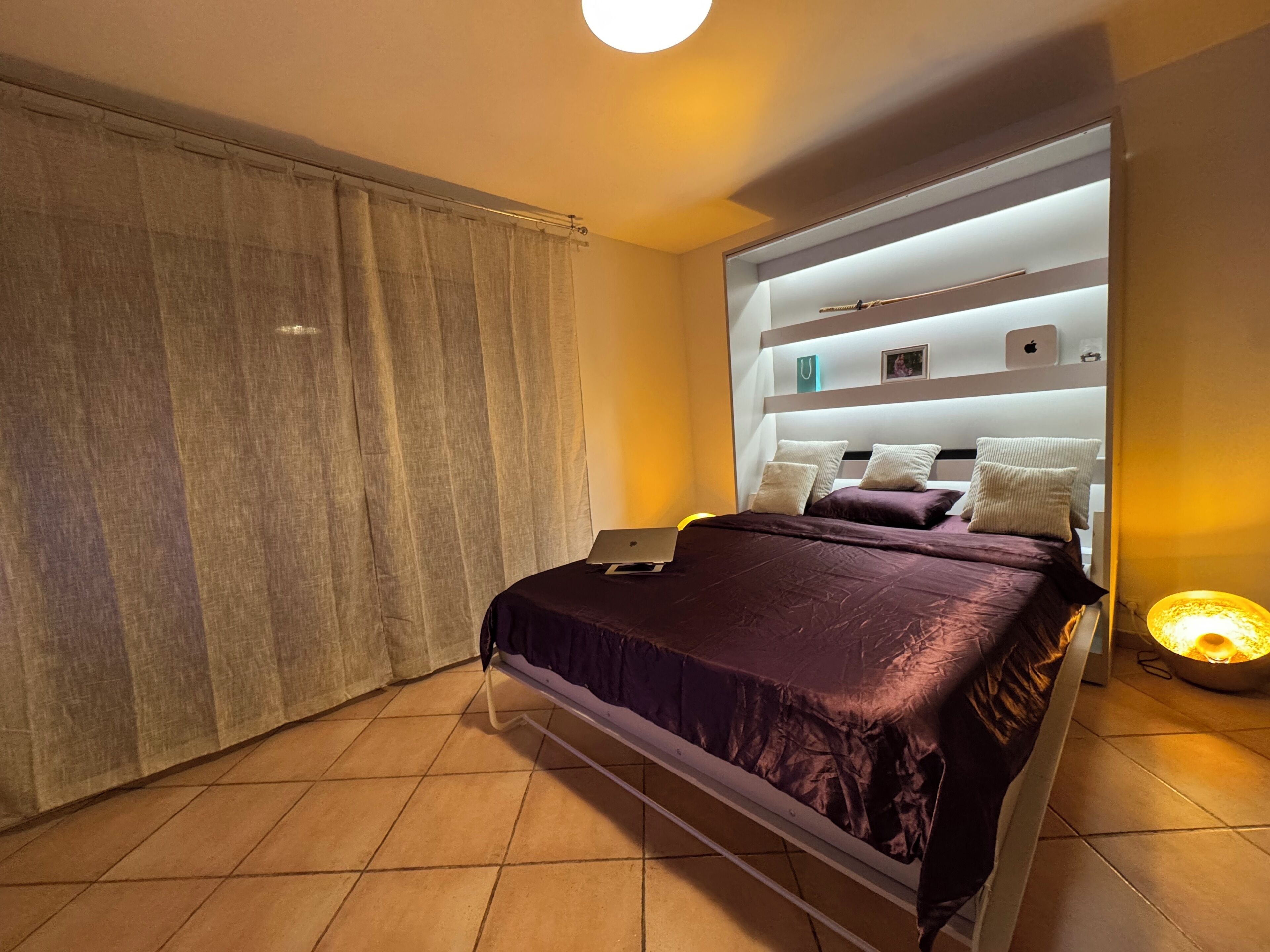 Un dormitorio moderno con cama, una computadora portátil sobre la cama, una lámpara de noche y una cortina.