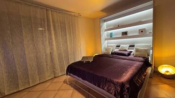 Un dormitorio moderno con cama, una computadora portátil sobre la cama, una lámpara de noche y una cortina.