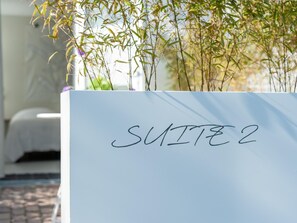 Ein weißes Schild mit der Aufschrift 'SUITE 2', platziert vor einem Bett mit weißem Kopfteil.