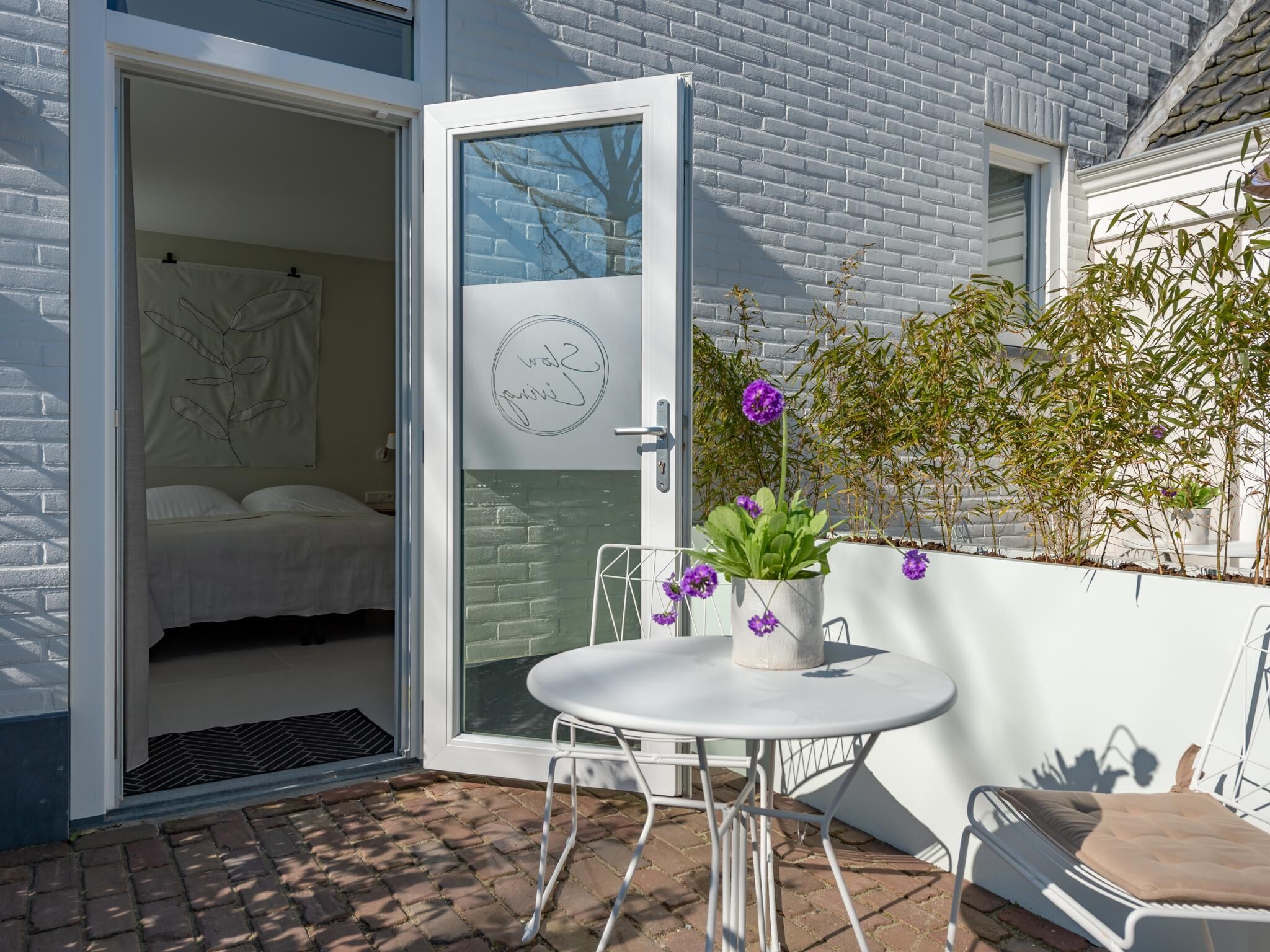 Een geopende deur die leidt naar een slaapkamer met een bed, een kleintje tafeltje met een bloempot en een stoel erbuiten.