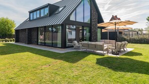 Ein modernes Haus mit großen Glasfenstern, einem geneigten Dach und einer Terrasse mit Gartenmöbeln.