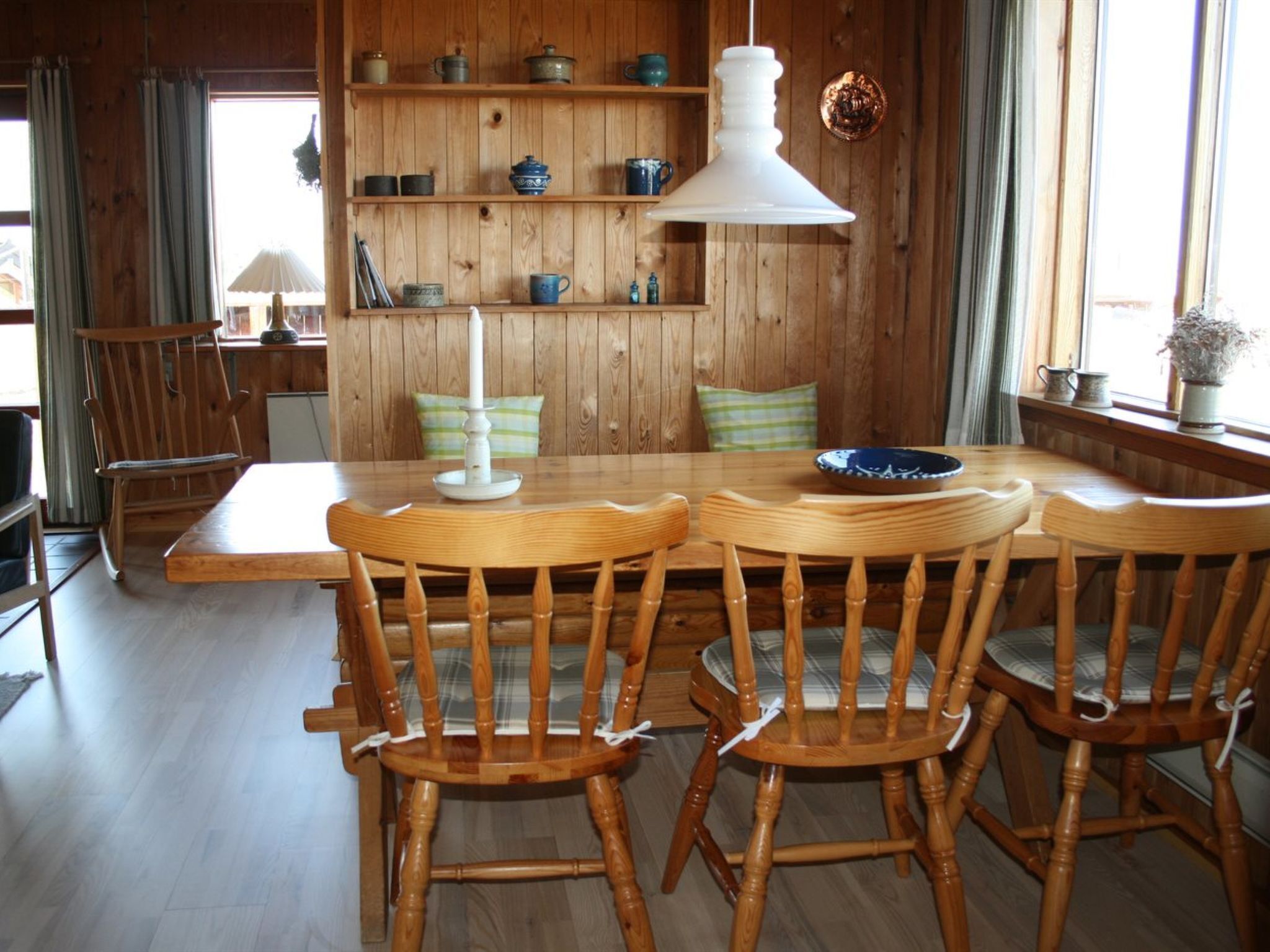 Uma zona de refeições de madeira com uma mesa, cadeiras, um candeeiro pendente e prateleiras com louças.