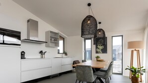 Une cuisine moderne avec des meubles blancs, une table à manger avec des chaises, des luminaires suspendus et une plante en pot.