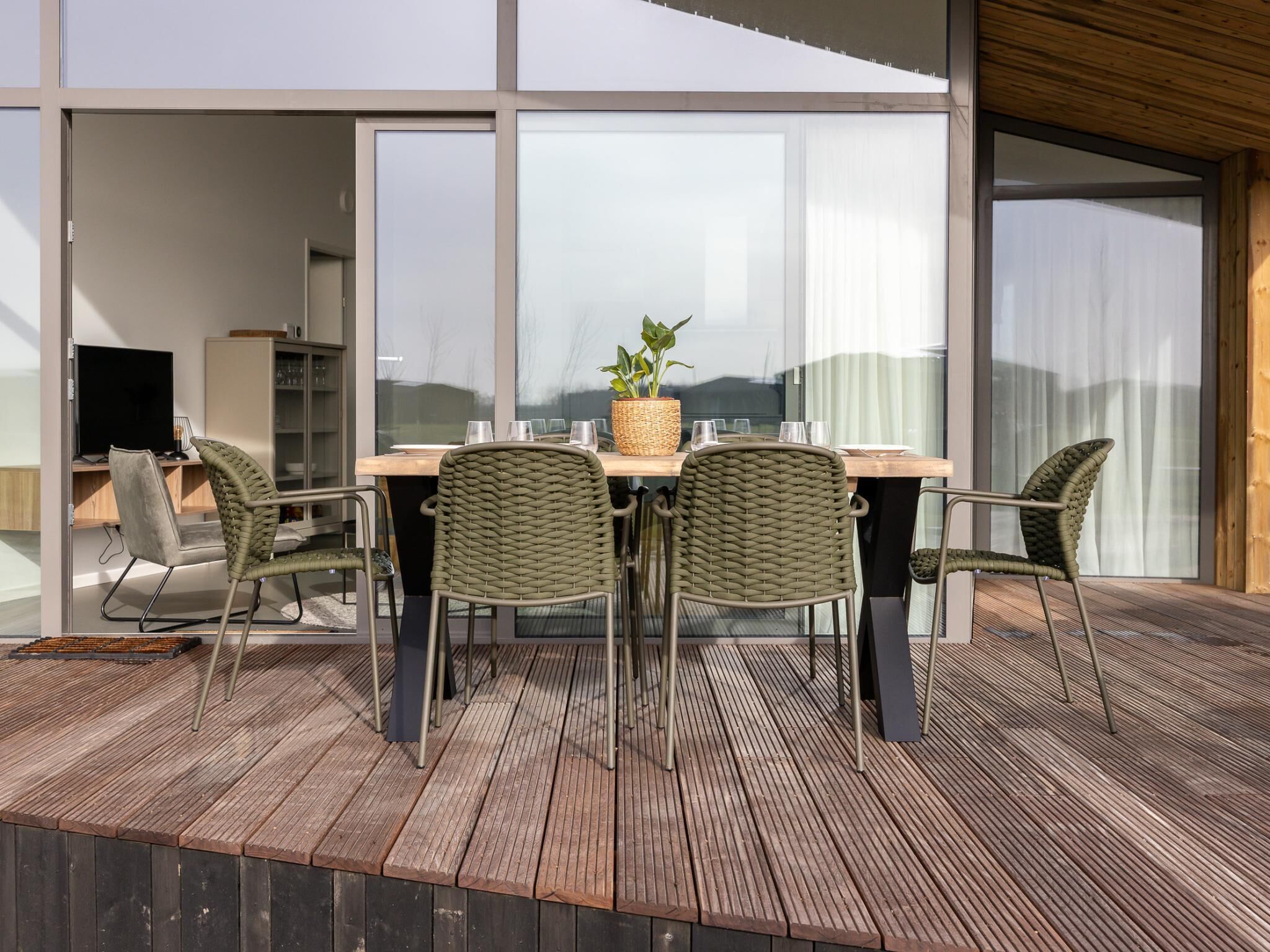 Uma área de refeições moderna ao ar livre, com mesa de madeira, cadeiras de vime, um vaso de planta e vista para a paisagem circundante.