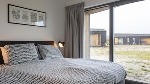 Une chambre à coucher avec un grand lit, une tête de lit, une lampe de chevet, une fenêtre avec des rideaux et un tableau encadré au mur.