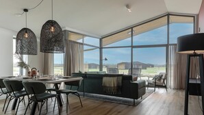 Uma sala de estar espaçosa com mesa de jantar, cadeiras, sofá e janelas amplas com vista para o exterior.