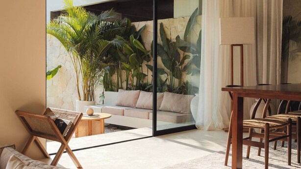 A modern living room with a sofa, a dining table, and chairs. There is a large window with a view of a garden. - Romantic 1BR Villa Canggu Pool & Sunset Balcony (Canggu)