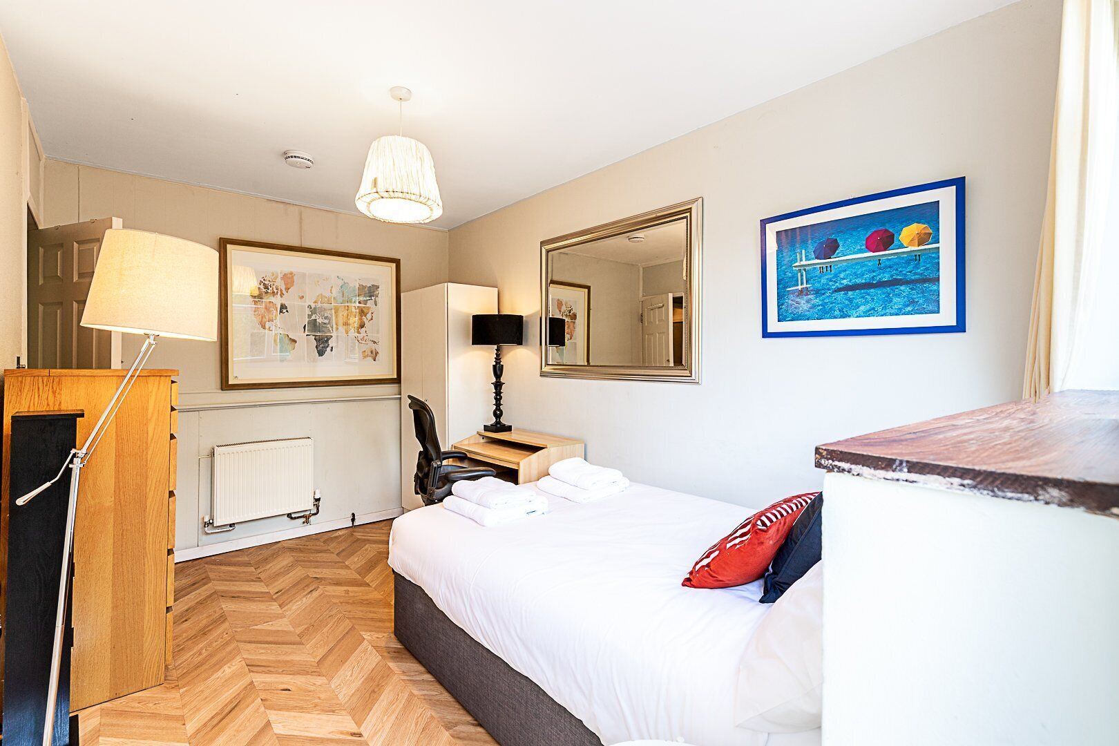 Ein Hotelzimmer mit Bett, Schreibtisch, Stuhl, Lampe, Heizkörper, Spiegel und einem gerahmten Bild an der Wand.