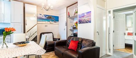 A compact living space with a dining area, a sofa, a chair, a table with a vase of flowers, and artwork on the walls.