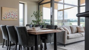 Un espace repas moderne avec une table en bois, des chaises à revêtement gris et une vue sur l’extérieur grâce à de grandes fenêtres.