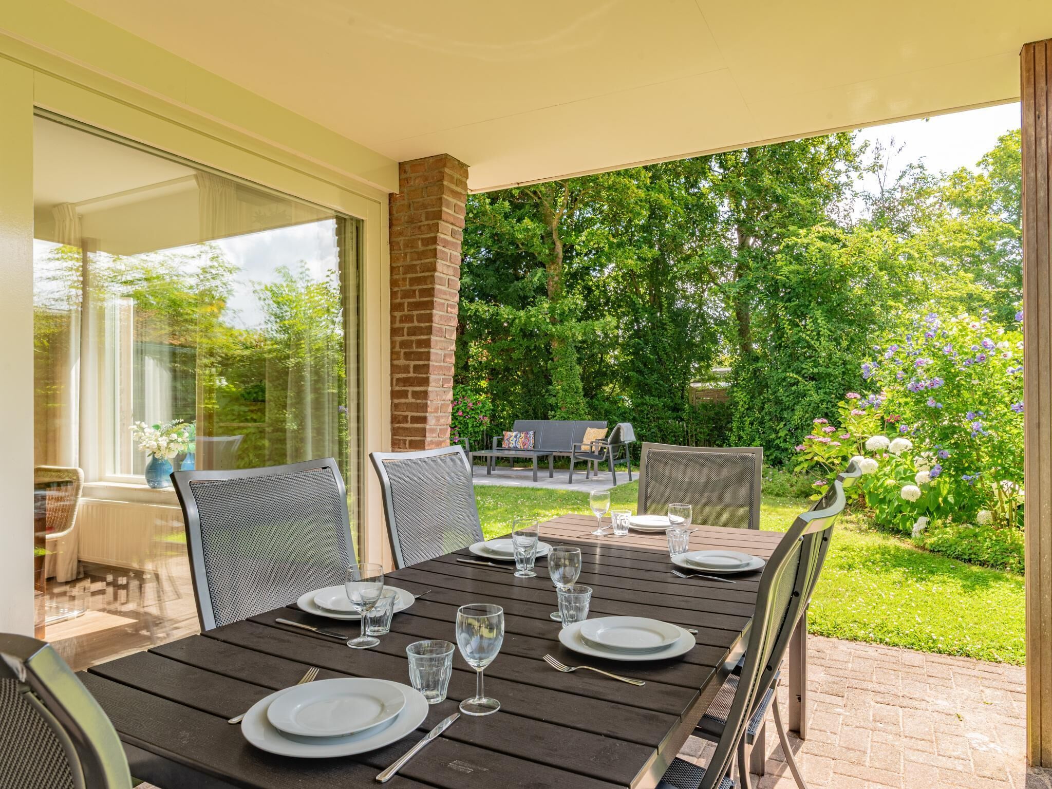 Eine überdachte Terrasse mit Esstisch, umgeben von Stühlen und Blick auf einen Garten mit Bäumen und Blumen.