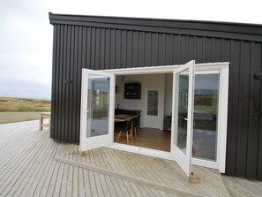 A modern building with a wooden deck, a dining area with chairs and a table, and a glass door leading inside.