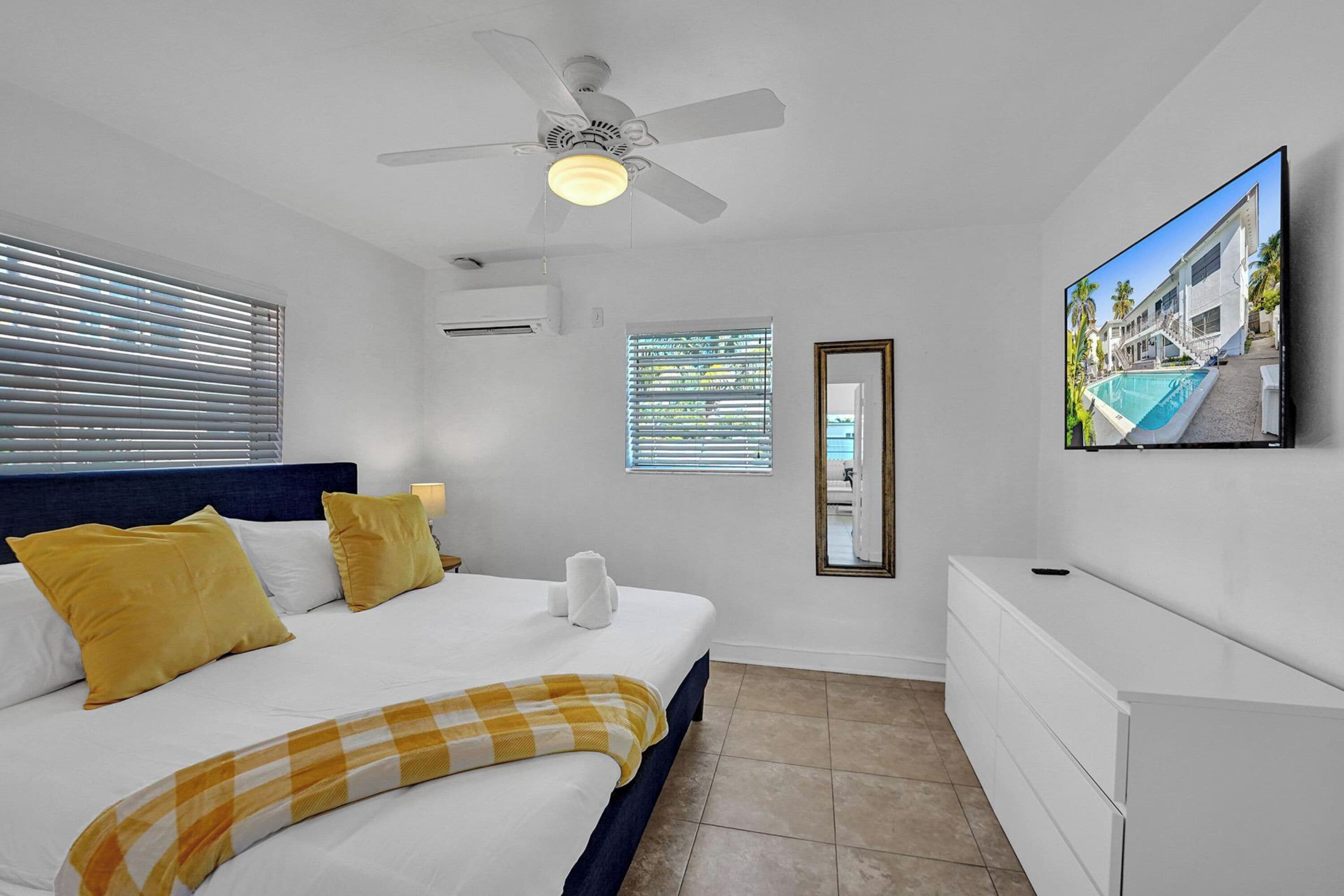 A bedroom with a bed, a ceiling fan, a TV, a window with blinds, and a door.