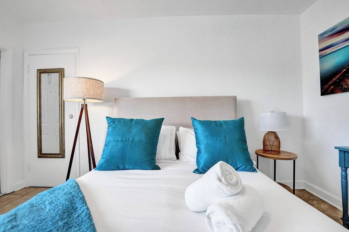 A hotel room with a bed, pillows, a lamp, a side table, and a painting on the wall.