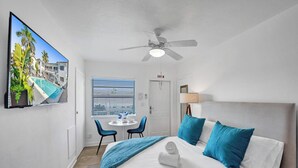 Habitación de hotel con cama, ventilador de techo, televisor que muestra vista a la piscina, ventana con persianas y puerta señalizada con 'EXIT'.