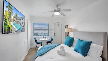 Habitación de hotel con cama, ventilador de techo, televisor que muestra vista a la piscina, ventana con persianas y puerta señalizada con 'EXIT'.