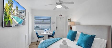 Habitación de hotel con cama, ventilador de techo, televisor que muestra vista a la piscina, ventana con persianas y puerta señalizada con 'EXIT'.