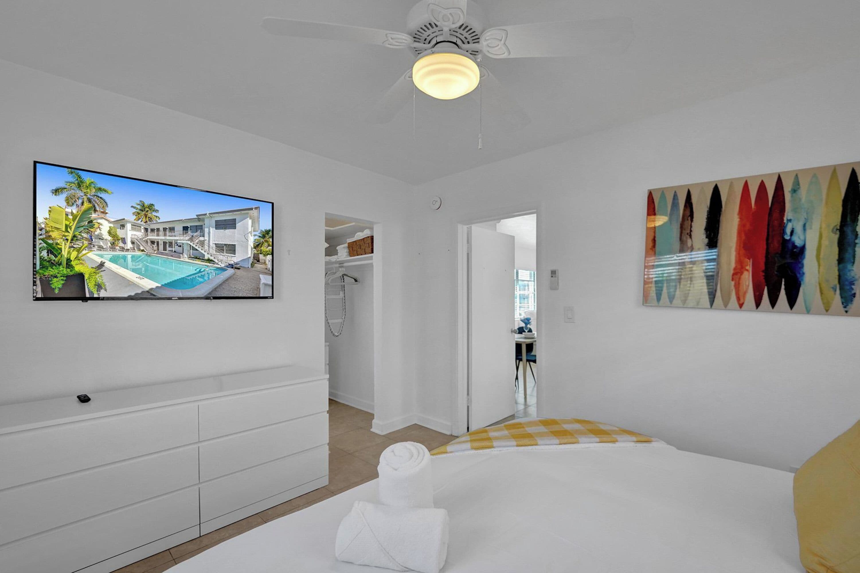 Ein Hotelzimmer mit Bett, Fernseher der auf einen Pool blickt, Deckenventilator und einem Gemälde.