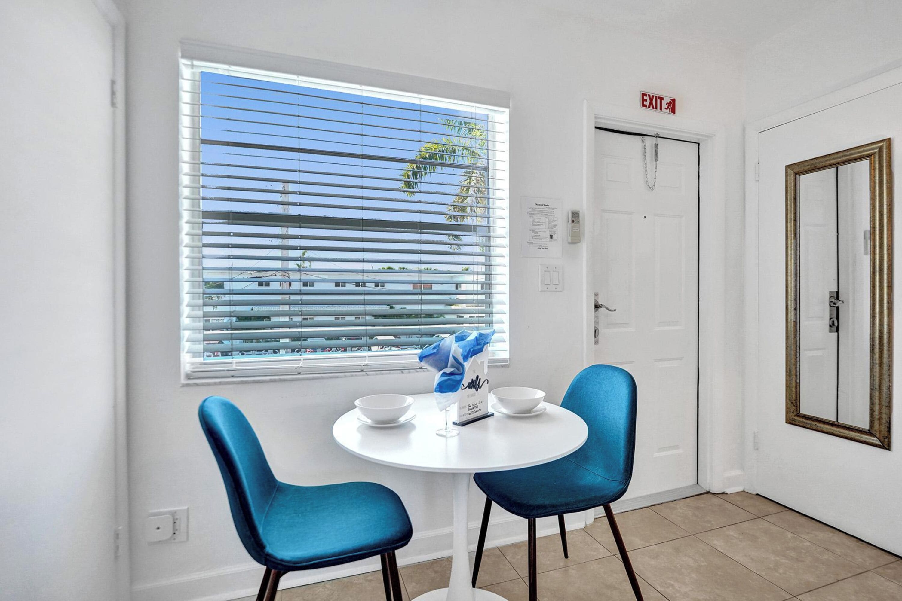 Una habitación con una mesa redonda, dos sillas azules, un ventanal con persianas, una señal de salida y una puerta con espejo.