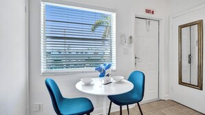 Una habitación con una mesa redonda, dos sillas azules, un ventanal con persianas, una señal de salida y una puerta con espejo.