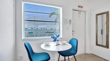 Una habitación con una mesa redonda, dos sillas azules, un ventanal con persianas, una señal de salida y una puerta con espejo.