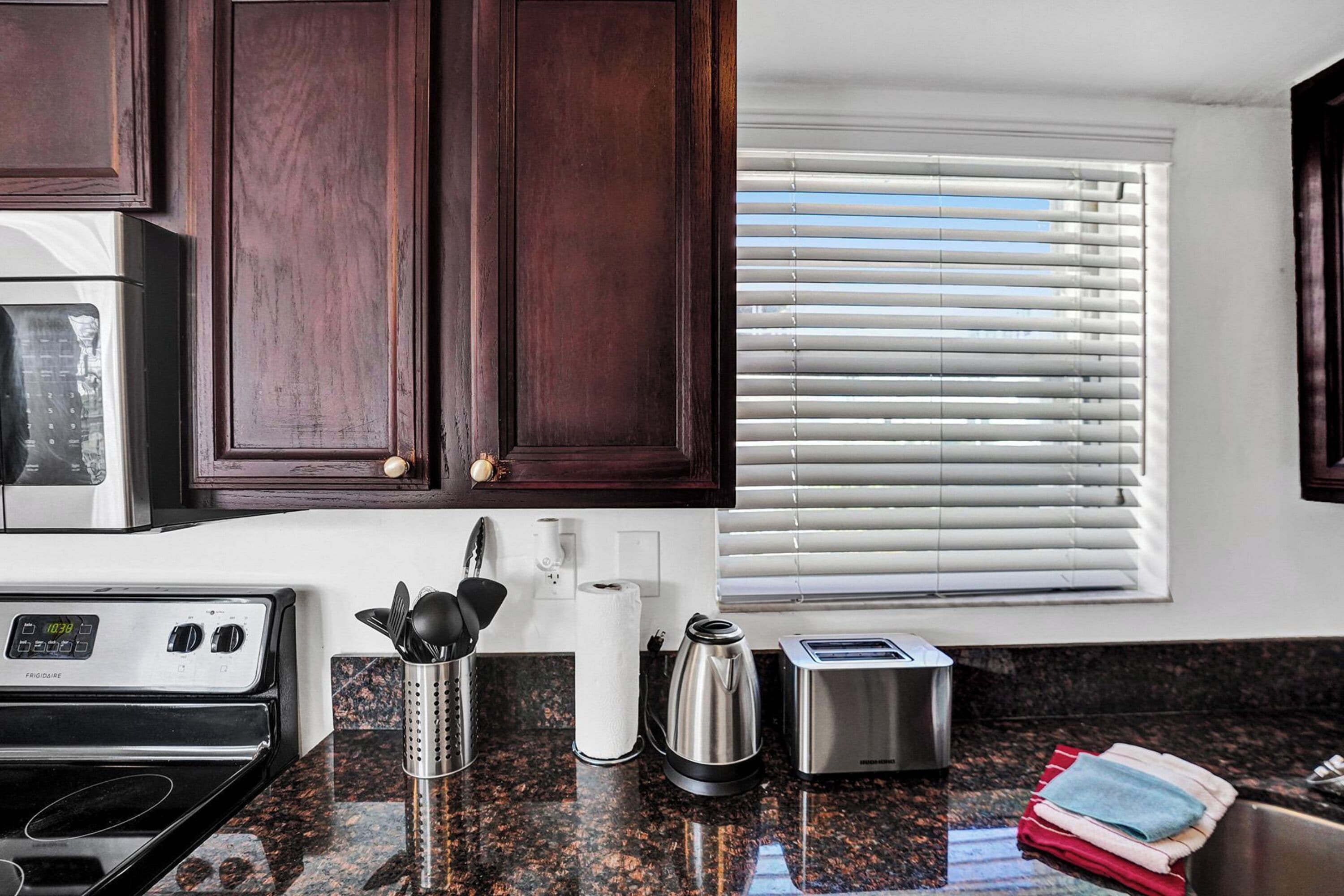 A kitchen with dark wood cabinets, a stainless steel refrigerator, a stove, a coffee maker, a toaster, and a window with blinds.