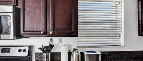 A kitchen with dark wood cabinets, a stainless steel refrigerator, a stove, a coffee maker, a toaster, and a window with blinds.