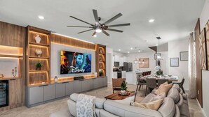 A modern living room with a flat-screen TV, a ceiling fan, and a dining area with a kitchen in the background. - Themed 7BR Villa | Lion King · Frozen · Minions | Mario Game Room | Pool & Spa (Clermont)