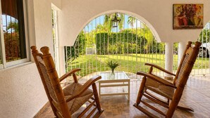 A covered patio with two rocking chairs, a glass table, and a view of a well-maintained garden. - Painted house- 4 pax, 4 min from the beach (Las Galeras)
