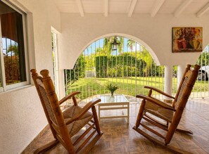 A covered patio with two rocking chairs, a glass table, and a view of a well-maintained garden. - Painted house- 4 pax, 4 min from the beach (Las Galeras)