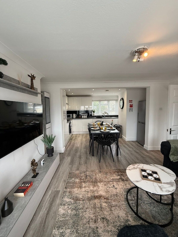 Une cuisine moderne avec des meubles blancs, un coin repas comprenant une table et des chaises, un téléviseur fixé au mur et une petite plante en pot posée sur le comptoir.