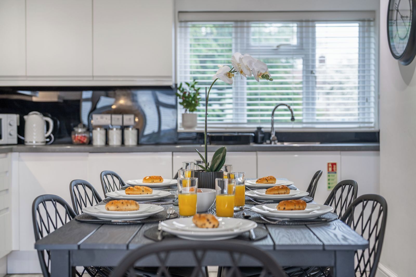 Eine moderne Küche mit weißen Schränken, einem Esstisch, der für das Frühstück gedeckt ist, mit Tellern, Gläsern und einer Orchidee im Topf.