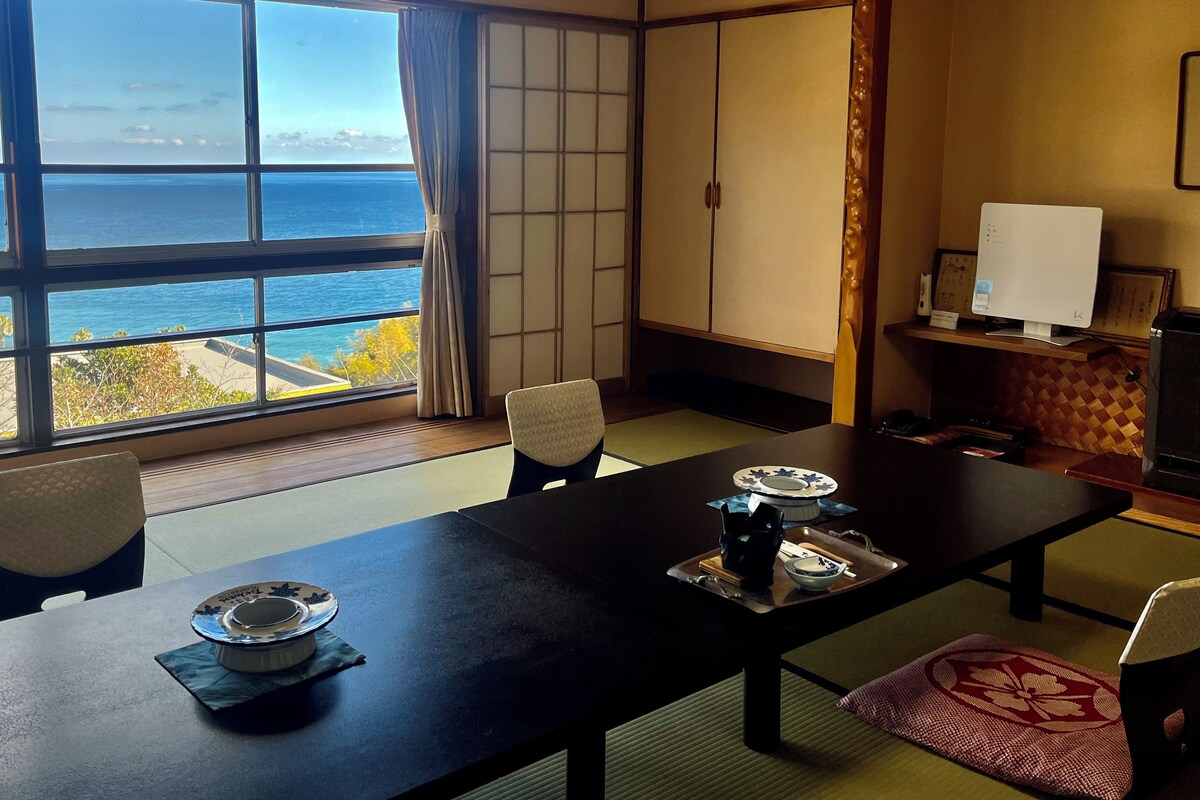 Ein traditioneller japanischer Raum mit Tatami-Böden, einem niedrigen Tisch mit einem Teeservice und Meerblick durch große Fenster.