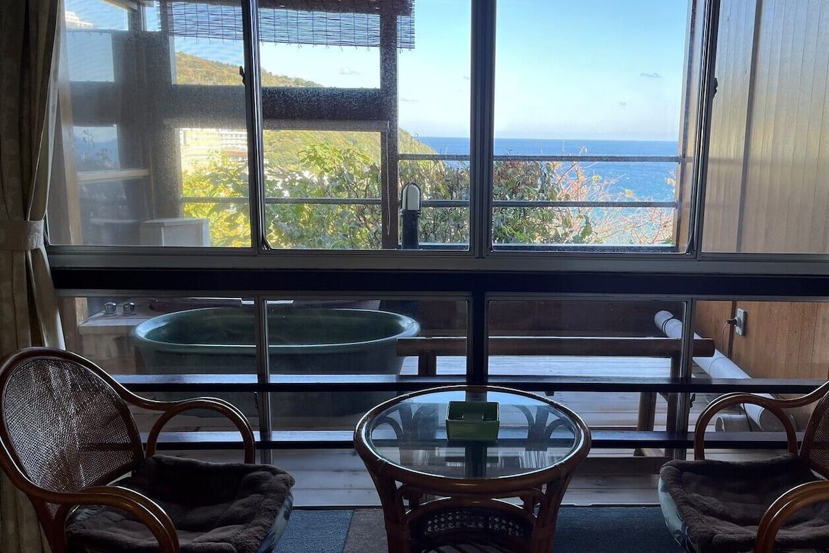 Een kamer met een groot raam, twee rieten stoelen, een kleine ronde tafel en uitzicht op de oceaan.