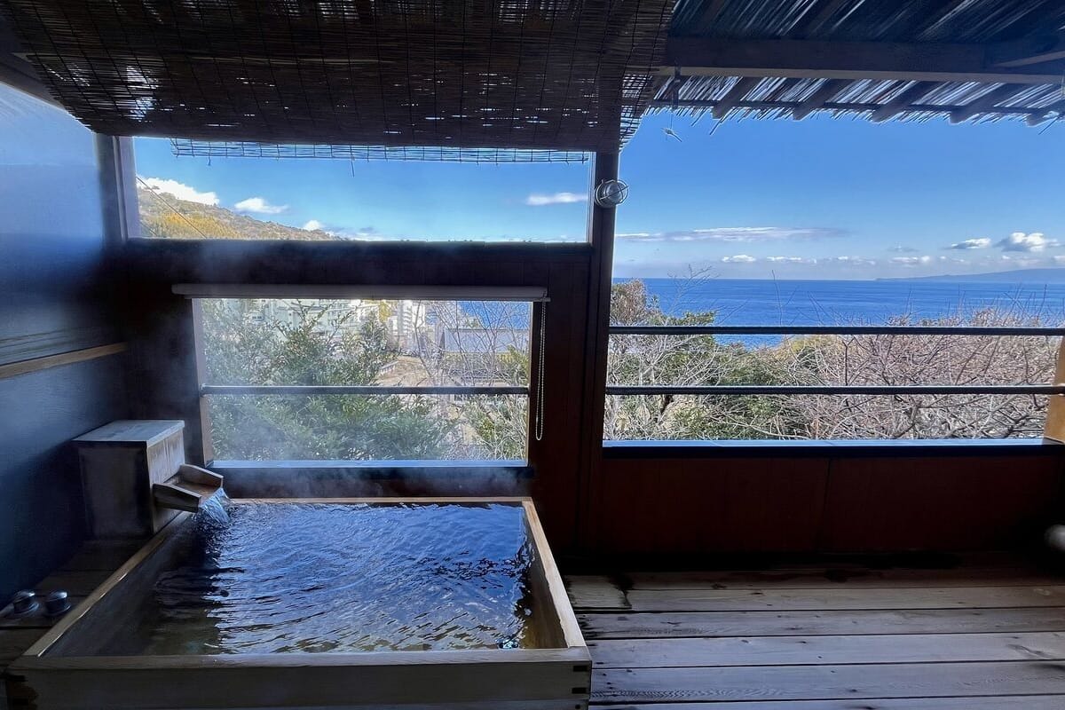 Een kamer met een groot raam dat uitkijkt over een bergachtig landschap en de zee, een houten vloer en een metalen tafel met een vloeistofoppervlak.