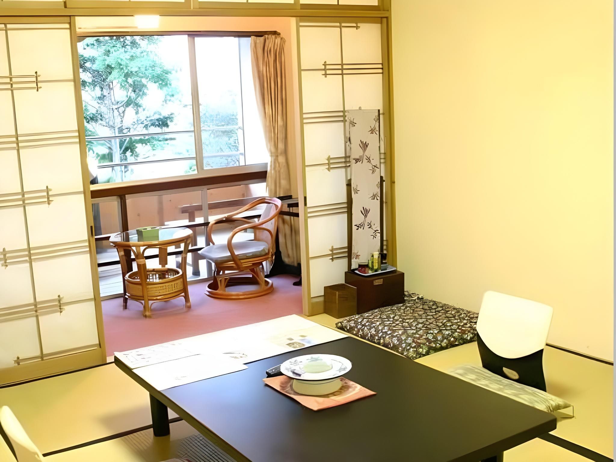 A traditional Japanese room with tatami flooring, a low table with a plate and chopsticks, and a sliding door leading to a balcony with a view of trees.