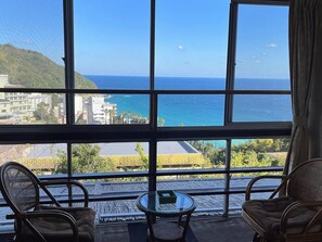Ein Zimmer mit großem Fenster und Blick auf die Küste, zwei Korbstühle und ein kleiner runder Tisch.