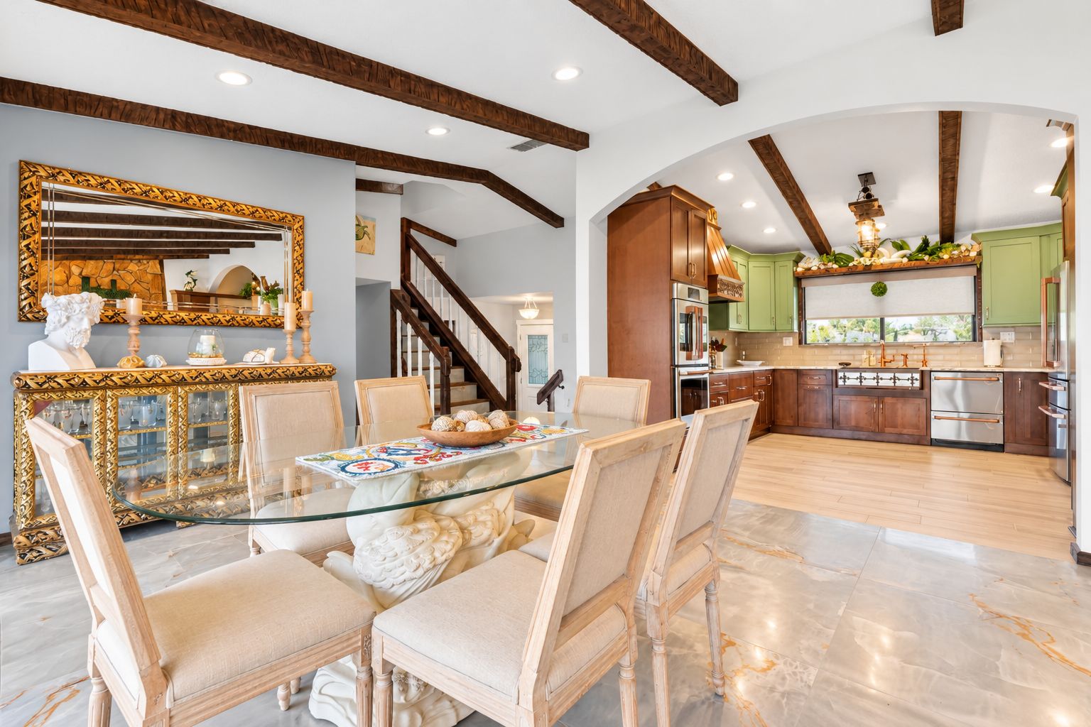 A spacious dining area with a glass table and chairs, a decorative console table with a bust, and a staircase leading to another level.