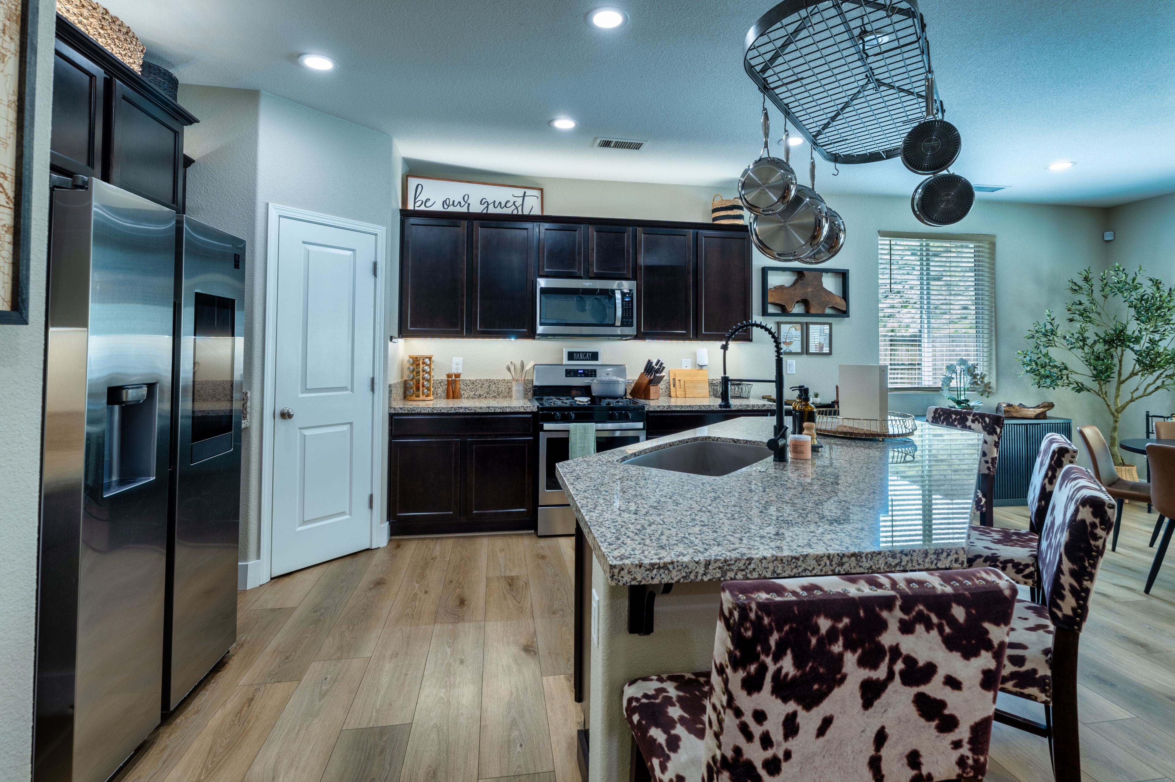 A modern kitchen with dark cabinets, stainless steel appliances, a granite countertop island, and a 'be our guest' sign.
