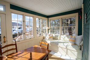 Una habitación acogedora con una mesa y sillas de madera, un banco con cojines y ventanas amplias con vistas a árboles cubiertos de nieve.
