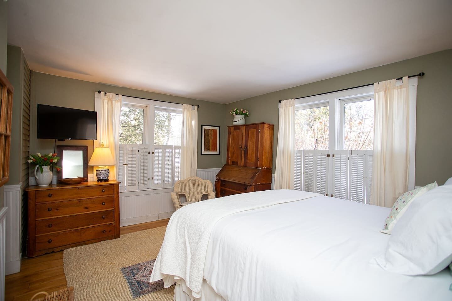 A bedroom with a bed, dresser, television, and windows with shutters.