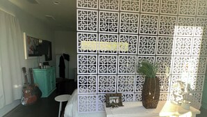 Una stanza con un letto, un tavolo consolle bianco con oggetti decorativi, una parete con motivo geometrico e un'anteprima di una zona cucina sullo sfondo.