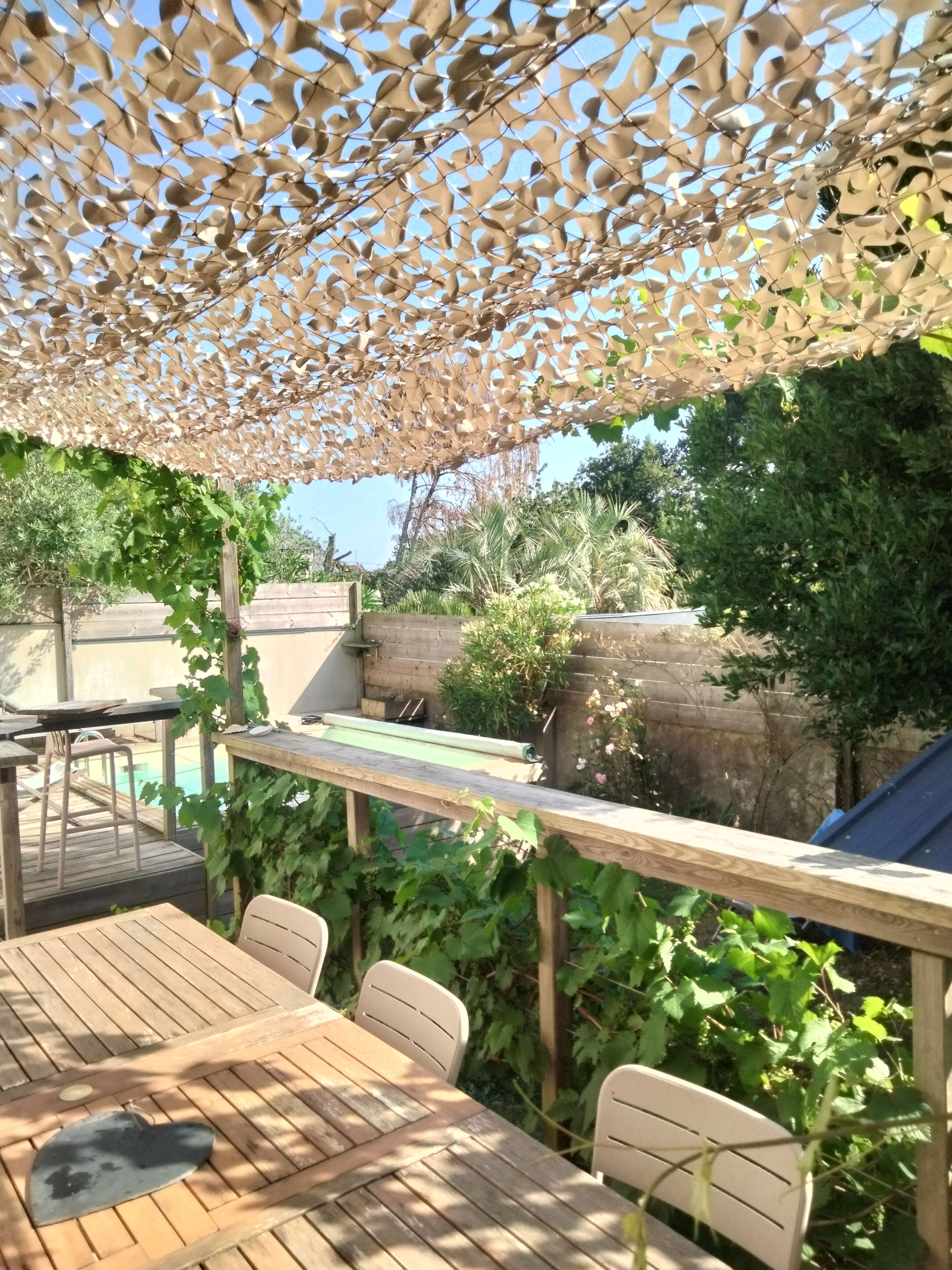 Une terrasse en bois avec une table et des chaises, une pergola ornée d’un motif évoquant des feuilles, et une piscine en arrière-plan.