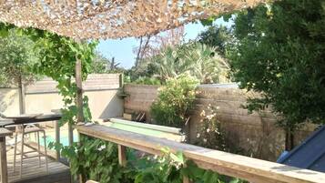 Une terrasse en bois avec une table et des chaises, une pergola ornée d’un motif évoquant des feuilles, et une piscine en arrière-plan.