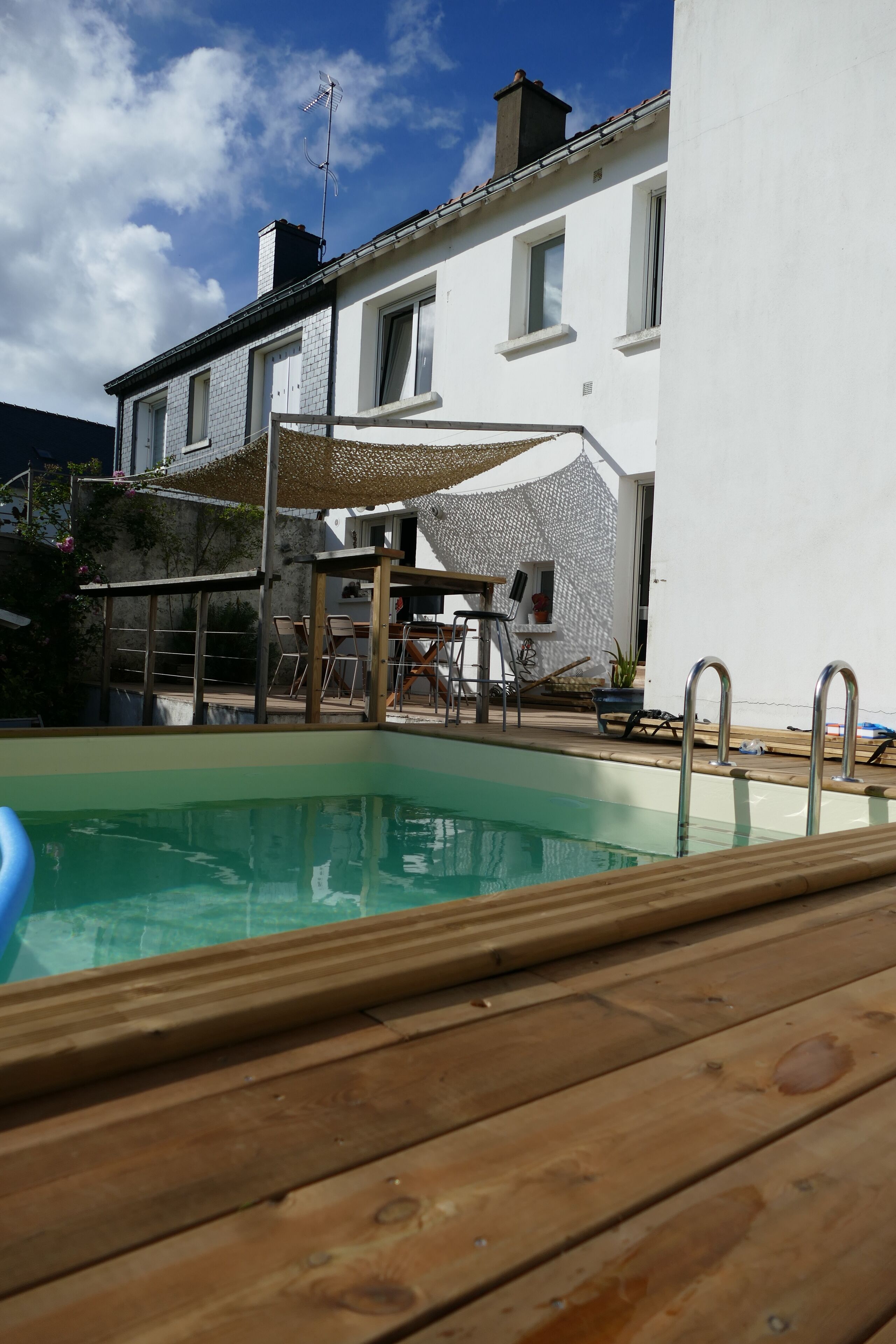 Une piscine avec une terrasse en bois, un bâtiment blanc doté de fenêtres et un espace extérieur aménagé et couvert.