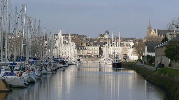 Ein Yachthafen mit zahlreichen Booten, die an einer ruhigen Wasserstraße liegen, umgeben von Gebäuden und einem markanten Kirchturm im Hintergrund.
