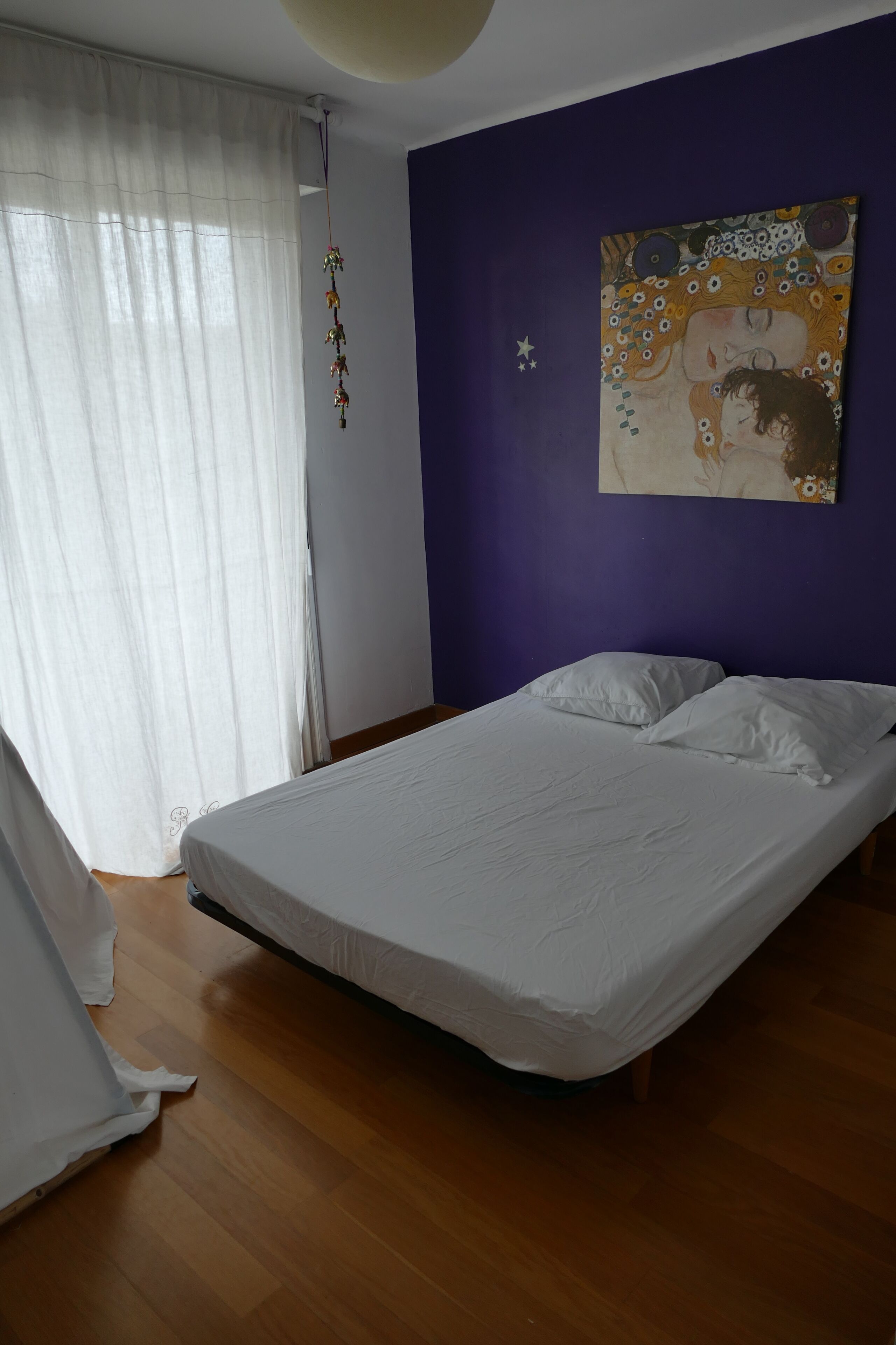 Une chambre à coucher avec un lit, des rideaux blancs, une décoration suspendue et un tableau au mur.